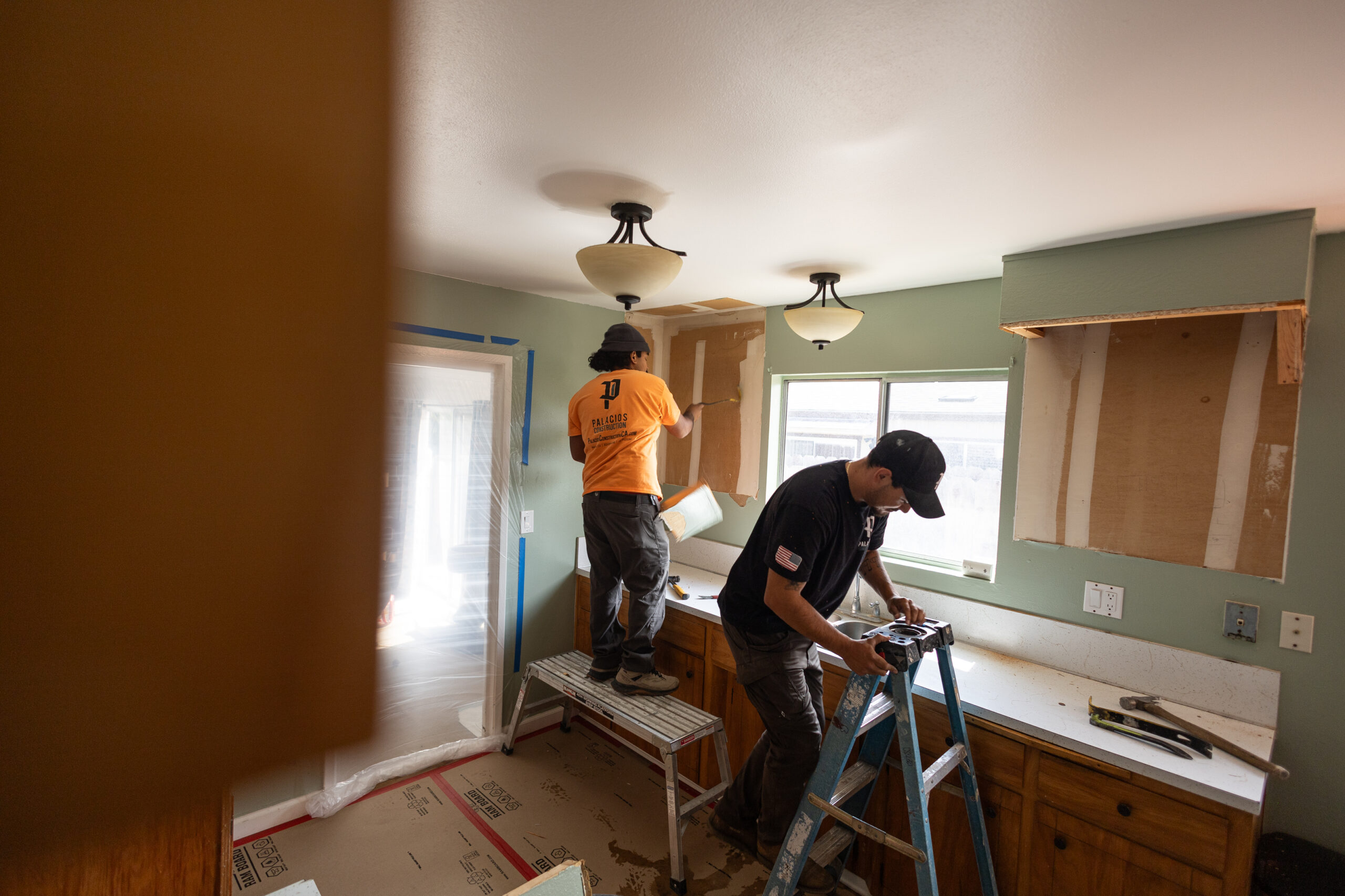 Workers finishing drywall and ceiling in a kitchen with protective plastic covering during an active.
